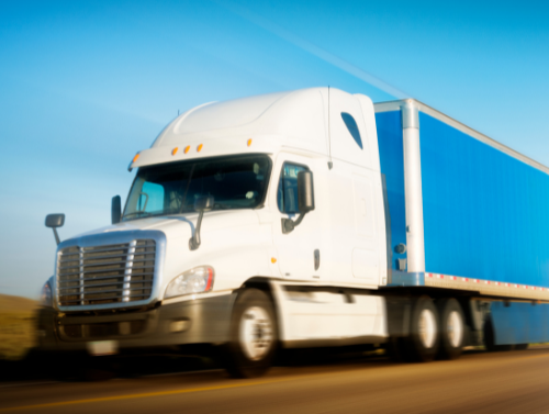 A white semi-truck with a blue trailer speeding on a highway under a clear sky.