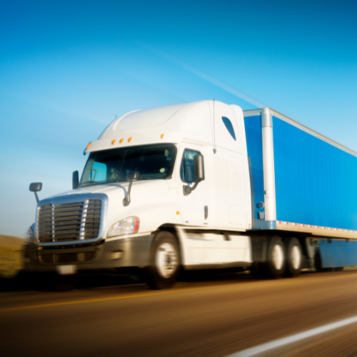 A white semi-truck with a blue trailer speeding on a highway under a clear sky.